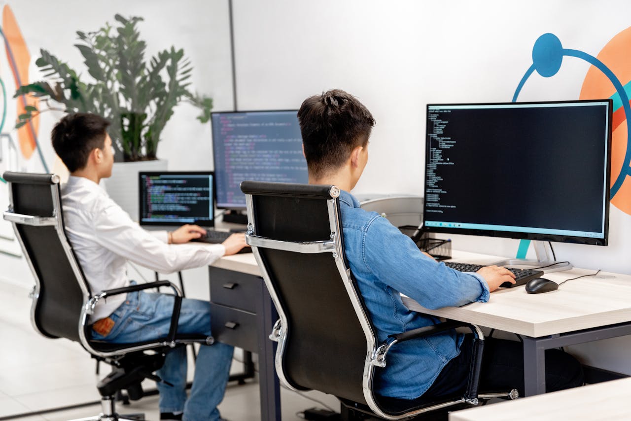 Two male developers at desks programming in a modern office workspace with large monitors.